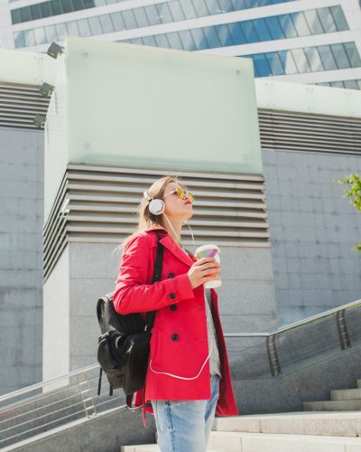 young hipster woman in pink coat, jeans in street with backpack and coffee listening to music on headphones, big city urban style trend, wearing sunglasses